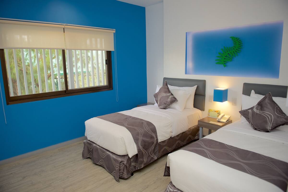 Een hotelkamer met twee bedden, een nachtkastje met een lamp, een wanddecoratie van een varen en een raam met jaloezieën.