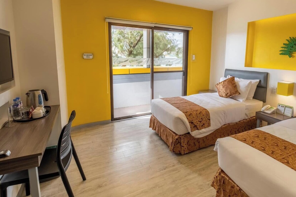 A hotel room with two beds, a desk with a kettle, a chair, a television, and a ceiling fan.