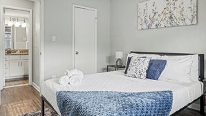 Un dormitorio con una cama, una mesita de noche, una lámpara fijada a la pared, una puerta y un baño visible a través de una puerta abierta.