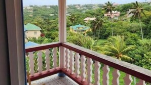 Un balcone con pavimento piastrellato e balaustra bianca, con vista su un paesaggio verde e rigoglioso con case e palme.