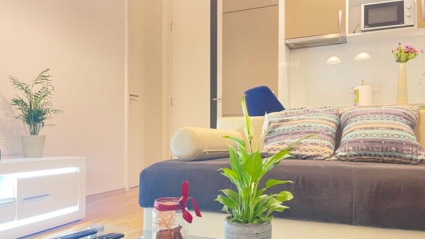 A modern living room with a sofa, decorative pillows, a glass coffee table, a potted plant, and a jar with dried flowers.