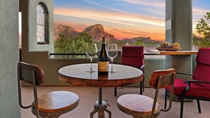Une table ronde en bois, sur laquelle sont posés une bouteille de vin et deux verres, entourée de chaises rouges, le tout placé devant un paysage pittoresque de montagnes et d’un coucher de soleil.
