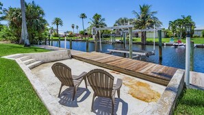 A concrete patio with two Adirondack chairs, a wooden dock extending into a body of water, and a gazebo structure. - Waterfront Fun with Heated Pool, Private Dock & Gulf Access – Tropical Delight - Roelens (Cape Coral)