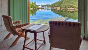 Une pièce avec vue sur une rivière paisible, des chaises en bois, une petite table et une terrasse en bois qui s’étend jusqu’à l’eau.