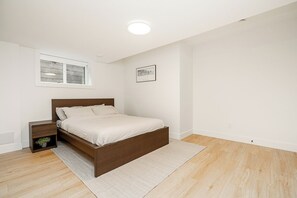 A bedroom with a wooden bed, a nightstand with a plant, a white wall, a window, and a framed picture. - Cozy & Spacious 2BR Guest Suite, Near Vancouver (Langley Township)