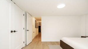 A bedroom with a bed, a wooden floor, a white door, and a hallway leading to another room. - Cozy & Spacious 2BR Guest Suite, Near Vancouver (Langley Township)