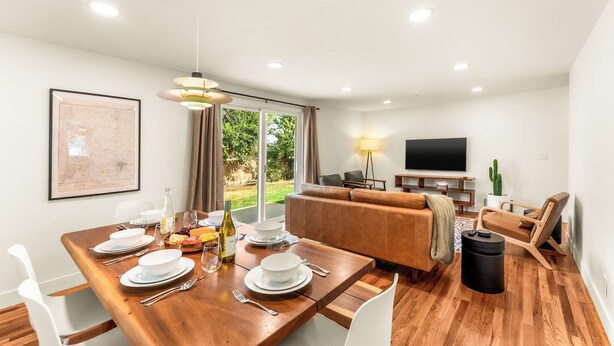 A modern living room with a dining area, a sofa, a TV, and a large window. - Modern Retreat w/ Hot Tub, Peloton, Gym, and BBQ (Westminster)