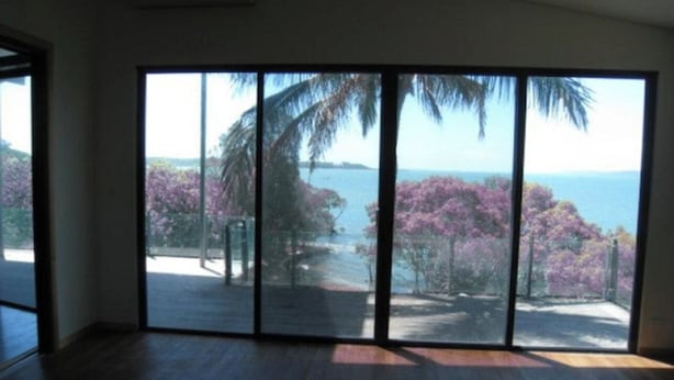 Quarto com uma grande porta de vidro deslizante que dá para uma varanda com vista para o mar e árvores.