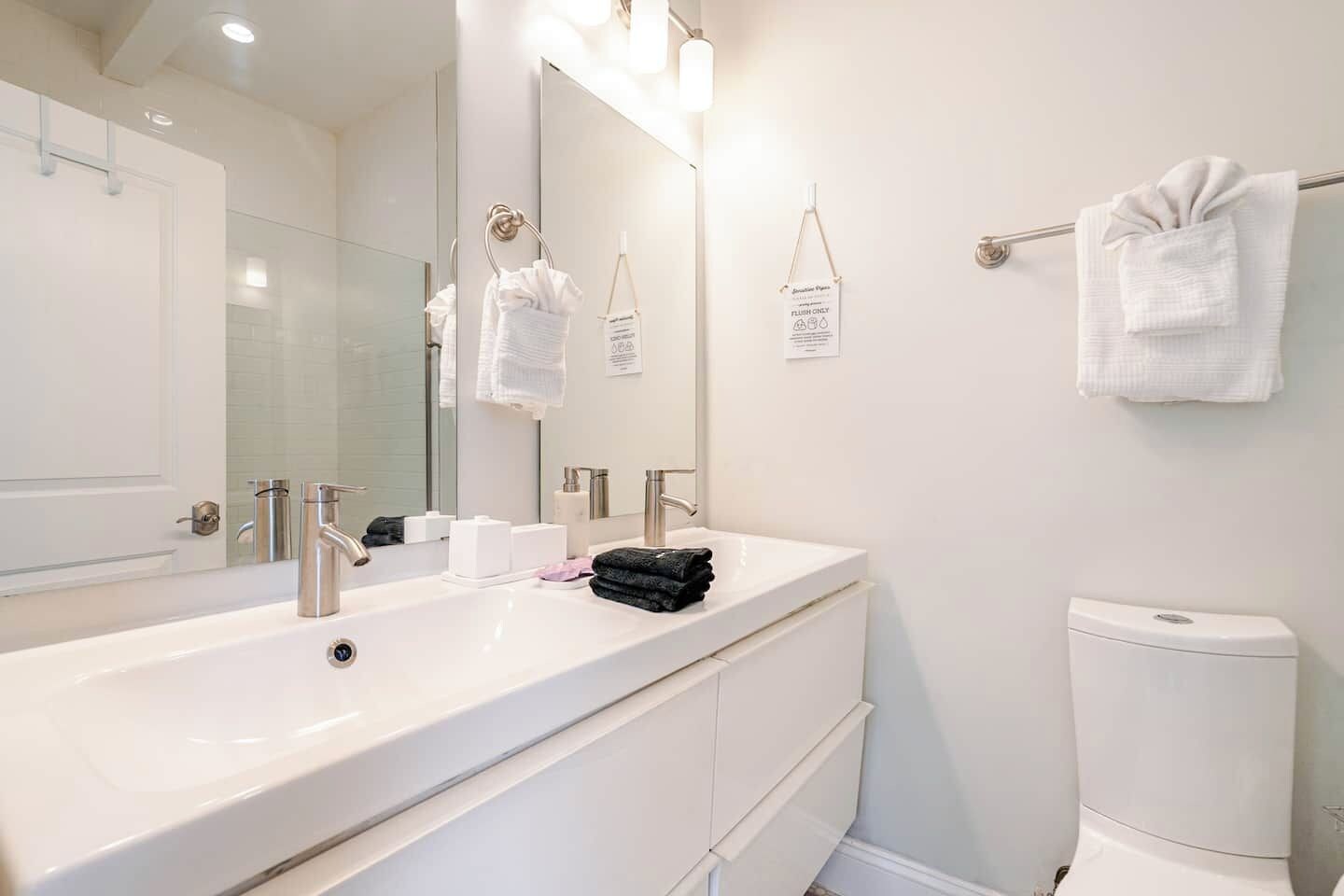 A bathroom with a white sink, two faucets, a mirror with lights, a toilet, and towels on a rack.