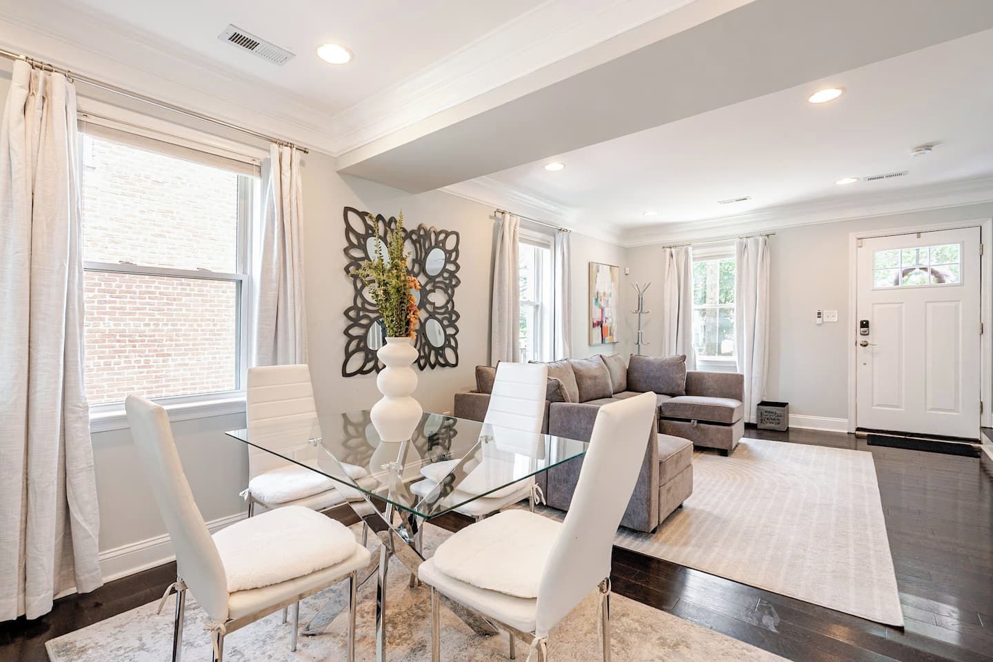 A modern living room with a glass dining table, white chairs, a grey sofa, and a large decorative mirror.