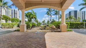 A covered outdoor area with a tiled floor, palm trees, and a view of high-rise buildings in the background. - Heated Pool, Sauna,| 3BR Condo| Waterside #835 (Fort Myers Beach)