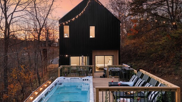 A modern two-story house with a hot tub on the deck, surrounded by trees and a sunset sky. - Pool + Hot Tub + Plunge | Scandinavian Wellness Retreat (Weaverville)