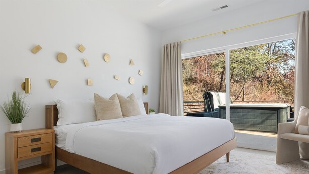 Quarto moderno com uma cama grande, uma mesa de cabeceira, uma planta em vaso e vista para uma varanda com móveis de exterior.
