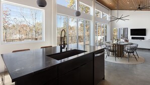 A modern kitchen with a large island, a dining area with a round table, and a large window overlooking a tree-lined view.