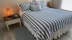 A neatly made bed with striped bedding, a bedside lamp, a small table with a tissue box, and a woven basket. - Charming 3-bedroom cottage in Seabrook, Texas (Seabrook)