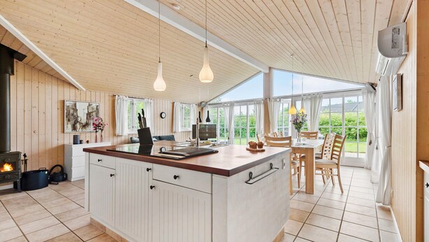 A modern kitchen with a large island, wooden ceiling, and large windows offering a view of the outdoors.