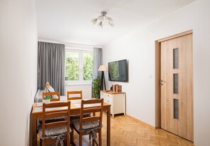 Een eetgedeelte met een houten tafel en stoelen, een raam met gordijnen, een plafondventilator, een klein kastje en een deur met een ruit.