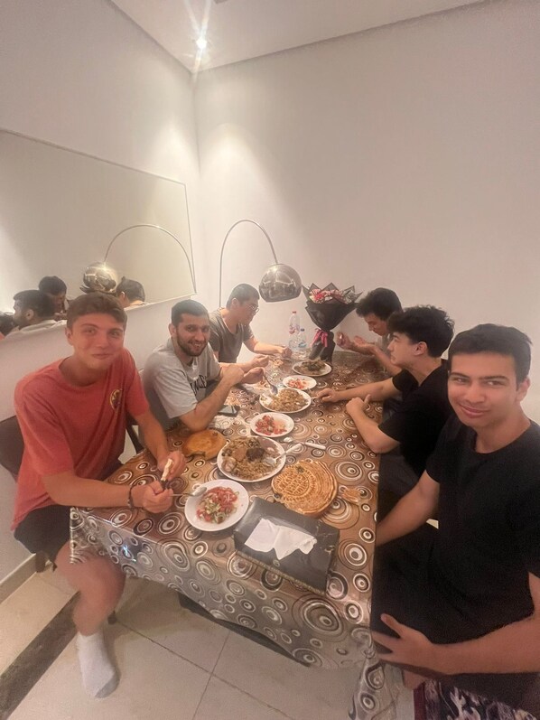 A group of people sitting around a table with various dishes, in a room with a patterned tablecloth and a mirror on the wall. - Bluewaters Islands Hostel (Dubai)