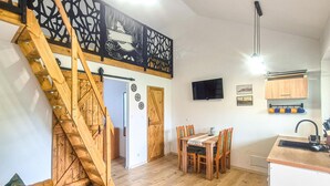 Une pièce spacieuse avec un escalier en bois, un coin repas avec des chaises en bois, une cuisine équipée d’un évier et de placards, et un téléviseur fixé au mur.