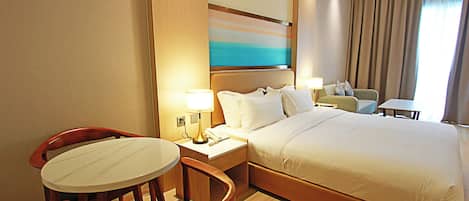 A hotel room with a large bed, a small table, a chair, a nightstand, and a painting on the wall.