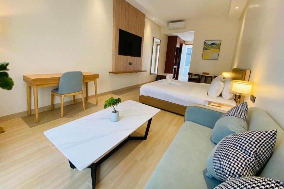 A modern hotel room with a bed, a desk, a sofa, a TV mounted on the wall, and a small plant on the table.