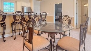 Un comedor con una mesa de vidrio y sillas ornamentadas, una cocina con armarios blancos y un acceso a un dormitorio.