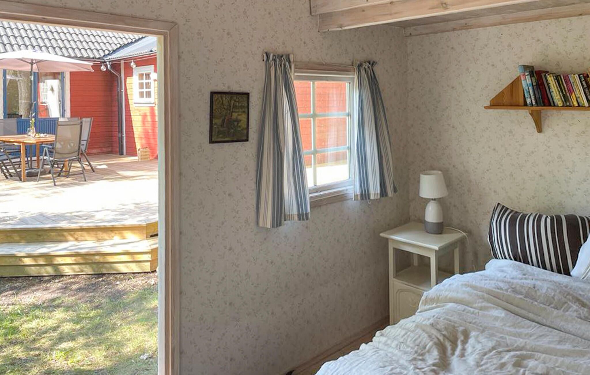 Une chambre à coucher avec un lit, une table de chevet avec une lampe, une fenêtre avec des rideaux, une bibliothèque et une vue sur une terrasse extérieure avec une table et des chaises.