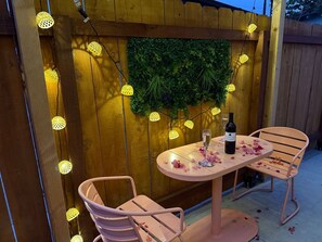 An outdoor patio with a round table and two chairs, a bottle of wine, and decorative lights.