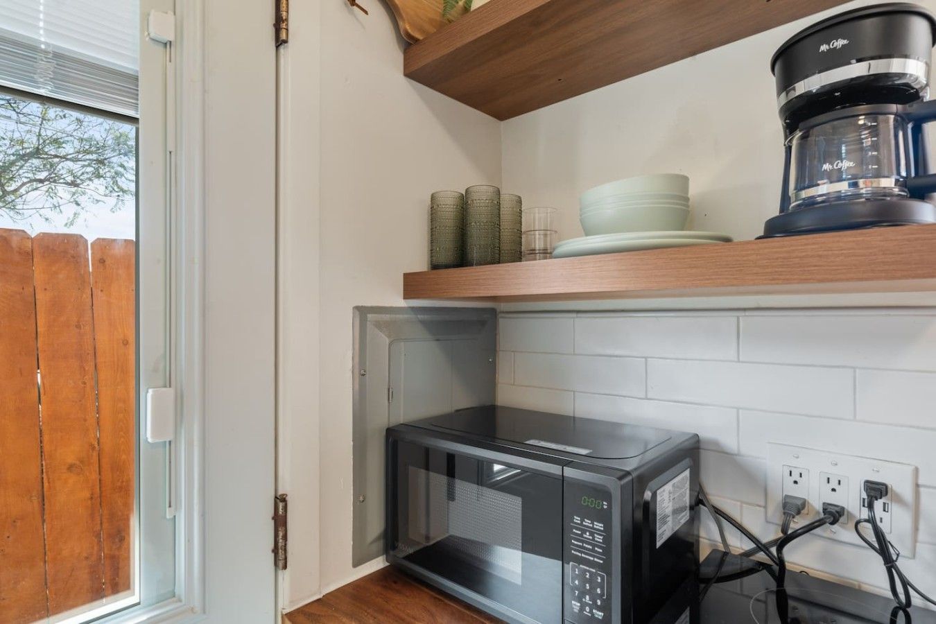 Una cocina con microondas, cafetera y estante de madera con vajilla.