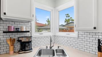 A kitchen with white cabinets, a double sink, a coffee maker, and a window with a view of houses.