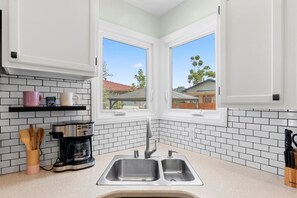 Cocina con alacenas blancas, un lavamanos doble, una cafetera y una ventana con vistas a casas.