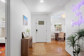 A hallway with a wooden floor, a white door, a small table with a plant, and a neon sign on the wall.