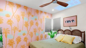 Une chambre à coucher avec un lit, un ventilateur de plafond, une fenêtre avec des stores et un mur orné d’un motif de palmiers.