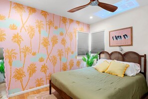 Een slaapkamer met een bed, een plafondventilator, een raam met zonwering en een muur met een palmboommotief.