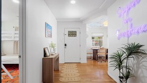 A hallway with a white door, a small table with a plant, and a neon sign on the wall.