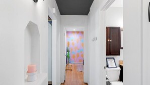 Un pasillo estrecho con pared blanca, suelo de madera, una vela rosa sobre un estante blanco, y un baño con inodoro y un certificado enmarcado en la pared.
