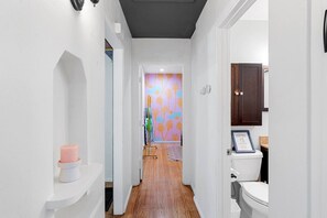 A narrow hallway with a white wall, a wooden floor, a pink candle on a white shelf, and a bathroom with a toilet and a framed certificate on the wall.