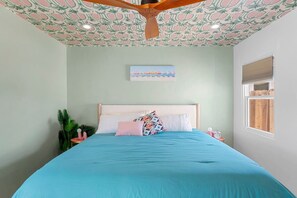 A bedroom with a bed, a ceiling fan, a window, and a wall-mounted artwork.