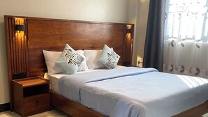 Une chambre à coucher avec une tête de lit en bois, un lit avec une literie blanche et des coussins à motifs léopard, une table de chevet, une lampe fixée au mur et une fenêtre avec des rideaux.