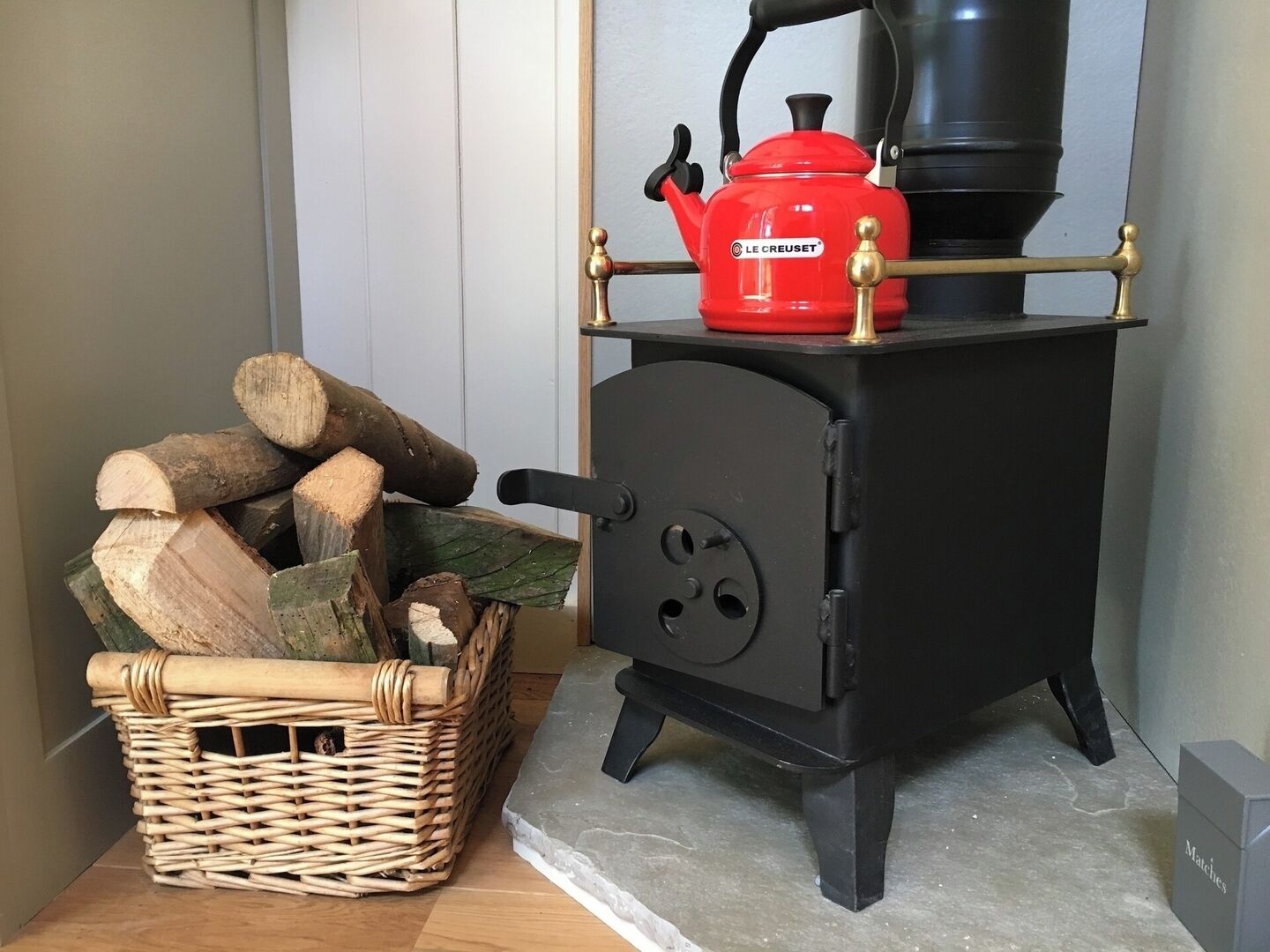 A black wood-burning stove with a red kettle on top, a wicker basket with logs, and a metal box labeled "Matches" on the floor.