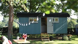 A small, single-story structure with a dark blue exterior and a corrugated metal roof. It features a door and windows, and is situated in a grassy area with trees and a small table with a vase of flowers.