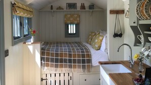 A compact bedroom with a bed, a window with curtains, a small window above the bed, a sink, and a mirror.