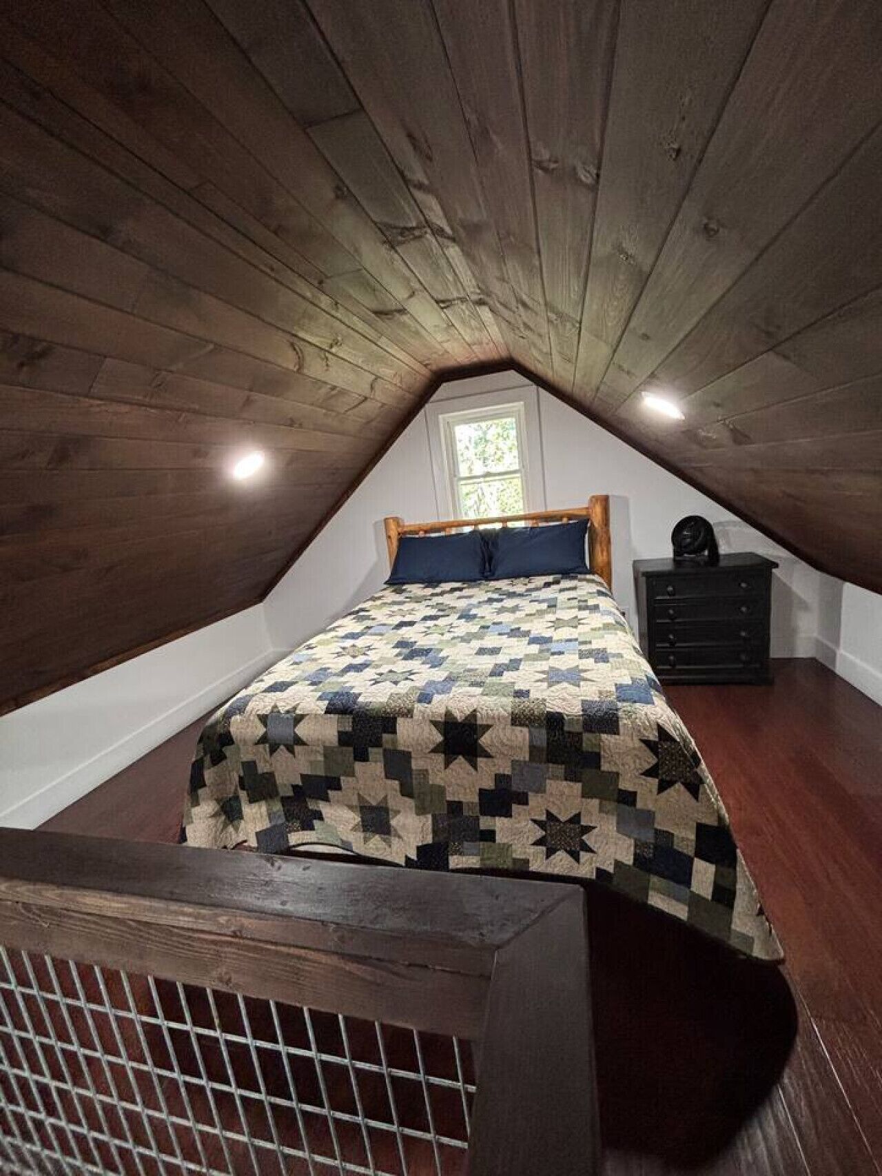 Um quarto de sótão pequeno com teto de madeira, uma cama com colcha, uma cômoda e uma janela.