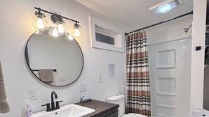 Un baño moderno con un espejo redondo, un lavamanos blanco, armarios de madera oscura, un inodoro blanco y una ducha con cortina de cuadros.