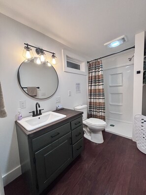 Un baño moderno con un espejo redondo, un lavamanos blanco, armarios de madera oscura, un inodoro blanco y una ducha con cortina de cuadros.