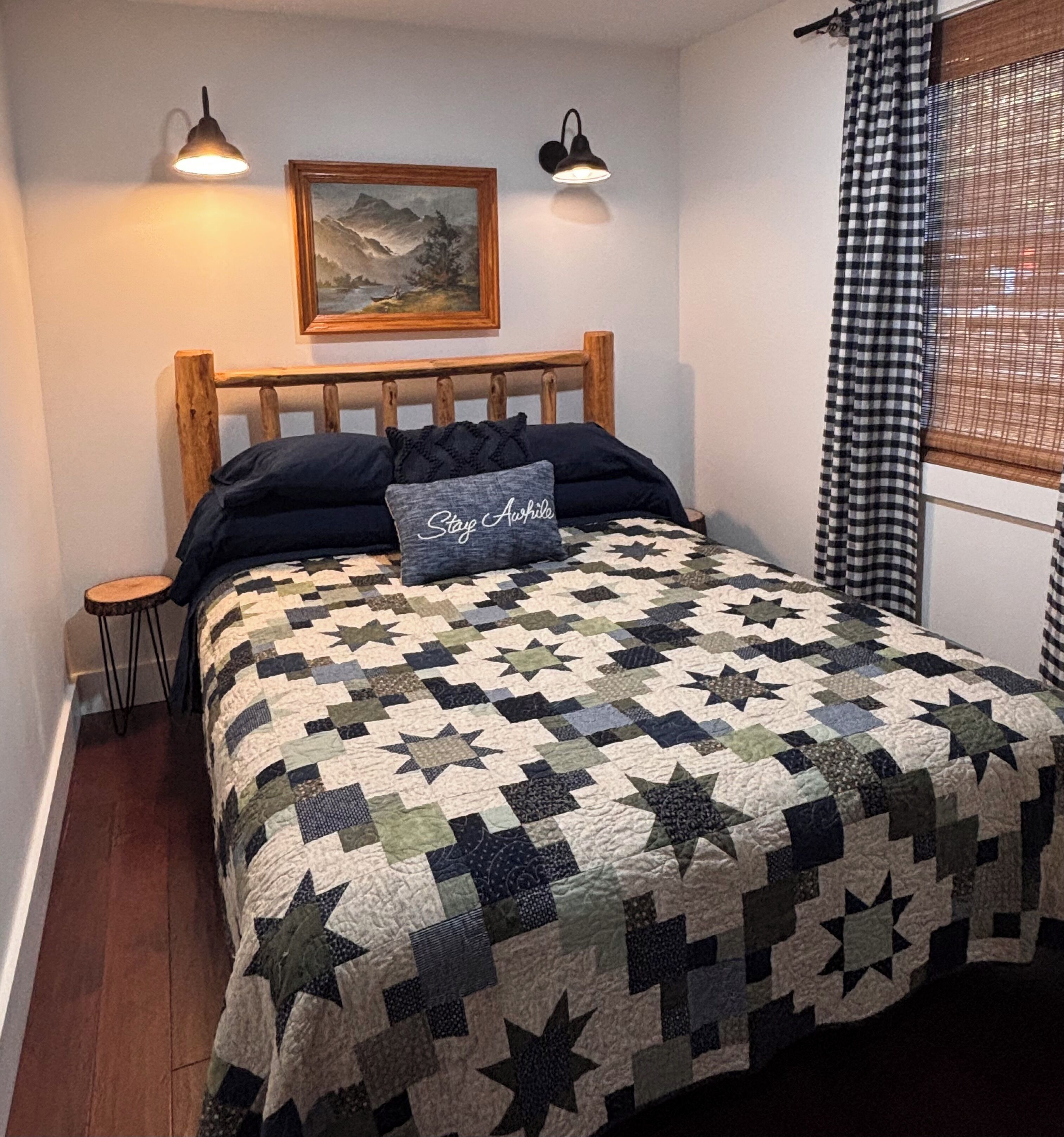A bedroom with a wooden headboard, a quilted bedspread, a small bedside table, a nightstand, a wall-mounted lamp, a framed picture, and a window with checkered curtains.