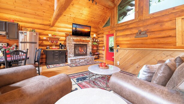 A cozy log cabin interior with a stone fireplace, wooden furniture, and a kitchen area.