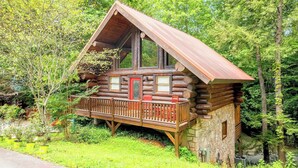 A log cabin with a wooden deck and a stone foundation, surrounded by lush greenery.