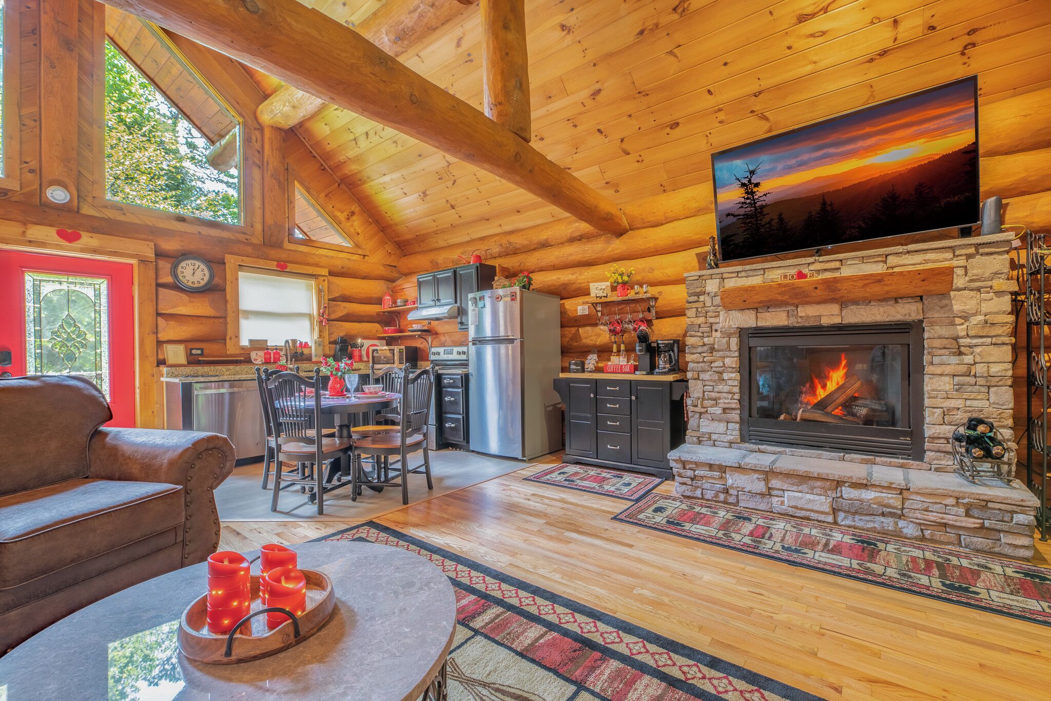 L’intérieur chaleureux d’un chalet en rondins, avec une cheminée en pierre, des poutres apparentes en bois au plafond, un coin repas et une cuisine.
