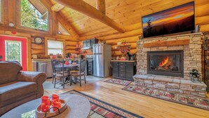 A cozy log cabin interior with a stone fireplace, wooden ceiling beams, a dining area, and a kitchen.